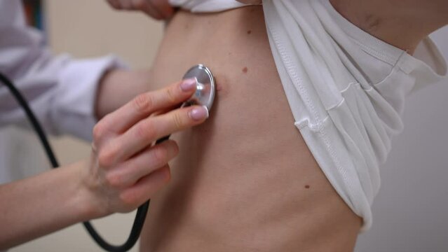 Close-up listening auscultation with stethoscope in pediatric clinic indoors. Unrecognizable Caucasian boy raising T-shirt as doctor using equipment diagnosing illness in hospital
