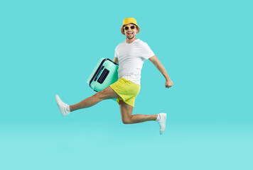 Fototapeta na wymiar Joyfully excited guy in summer clothes runs and jumps with suitcase on light blue background. Young man in shorts, panama and t-shirt in hurry summer vacation with crazy funny facial expression.
