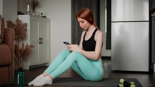 A girl in sportswear is sitting on a rug typing messages on her phone.