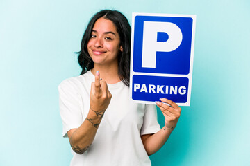 Young hispanic woman holding parking placard isolated on blue background pointing with finger at you as if inviting come closer.