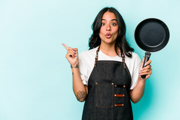 Young hispanic cooker woman holding frying pan isolated on blue background pointing to the side