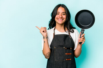 Young hispanic cooker woman holding frying pan isolated on blue background smiling and pointing...