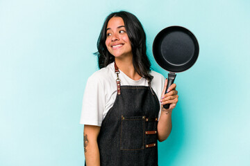 Young hispanic cooker woman holding frying pan isolated on blue background looks aside smiling, cheerful and pleasant.