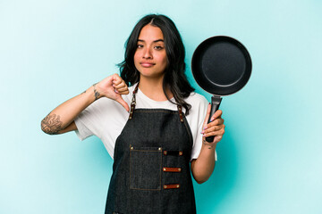 Young hispanic cooker woman holding frying pan isolated on blue background showing a dislike...