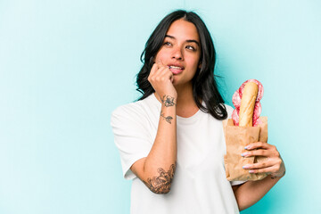 Young hispanic woman eating a sandwich isolated on blue background relaxed thinking about something looking at a copy space.