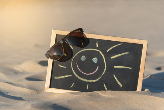 Pizarra negra con el marco de madera y unas gafas de sol en su esquina con un sol sonriente dibujado con tizas de colores sobre la arena clara de una playa y con el brillo del sol reflejado sobre ella