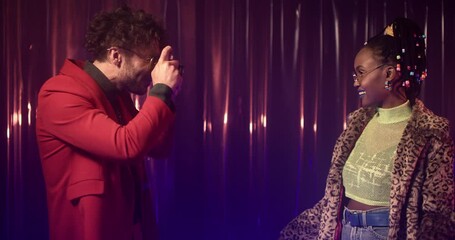 Smiling photographer in bright clothes takes picture of young woman in fur coat and sunglasses, showing peace signs with hands. Guy and girl spend evening together at night club.