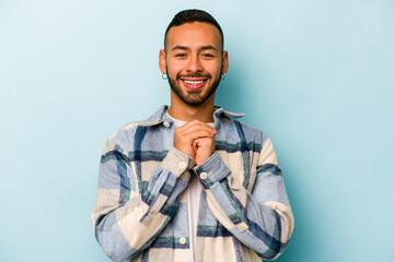 Young hispanic man isolated on blue background praying for luck, amazed and opening mouth looking to front.