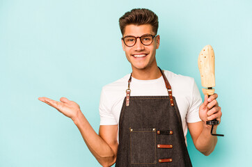 Young caucasian shoemaker isolated on blue background showing a copy space on a palm and holding another hand on waist.