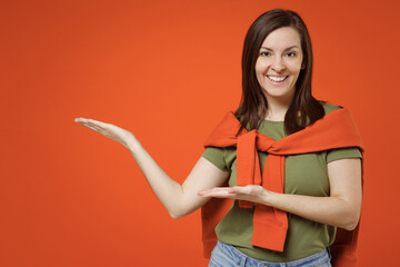 Young smiling fun happy woman 20s wearing khaki t-shirt tied sweater on shoulders point hands arms aside on workspace area mock up isolated on plain orange background studio. People lifestyle concept.