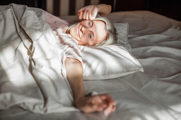 Happy middle aged woman sits on bed and stretching arms