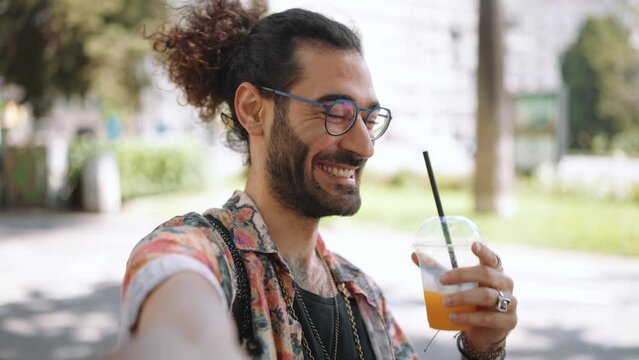 Positive curly-haired bearded man in eyeglasses drinking juice at the camera outdoors