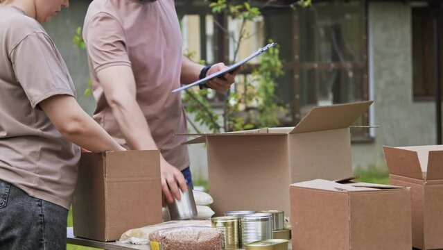 Group of volunteers putting grocery products, foodstuff to food donations box outdoors. People volunteering to sort donations for charity. Volunteers packs canned food and cereals in a boxes
