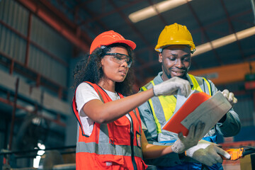 Two professional engineer/worker/technician use clipboard discuss work, walk in steel metal manufacture factory plant industry. Black African American man and woman wear hard hat check quality machine