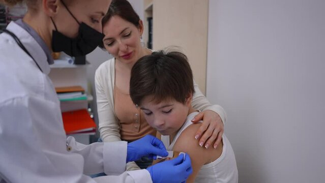 Live camera follows positive pediatrician walking to boy sticking medical patch on shoulder after vaccination. Smiling Caucasian patient thanking doctor for coronavirus vaccine injection