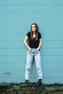 A teenage girl with long hair wearing black, denim and combat boots leaning against a blue cinder block wall and looking away with attitude