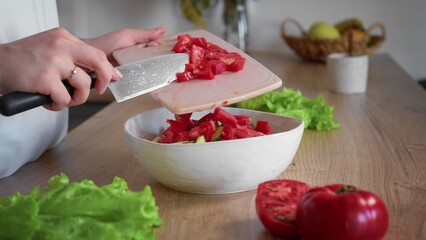 Close-up of an unrecognizable woman's hand cutting a tomato with a kitchen knife. The woman transfers the chopped tomato to a bowl. Cooking in the kitchen.