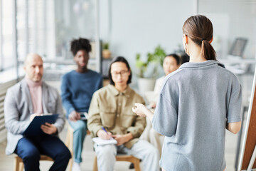 Back view of female coach giving lecture or seminar in office to diverse group of people, copy space