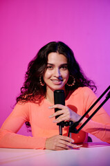 Vertical portrait of joyful female influencer sitting at desk in studio in pink neon light whispering in microphone for ASMR vlog