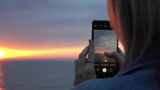Women's hands close-up hold smartphone and photograph on it beautiful orange sunset on sea. Clouds are colored red. Rear view. Festive mood on vacation.