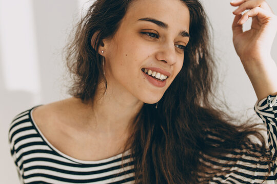 Close-up picture of beautiful young brunette female with ideal face features, wearing striped black and white blouse, looking aside smiling widely, long wavy hair falling on her shoulder