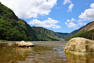 View across the water along Upper Lake in the Glendalough Valley in the Wicklow Mountains National Park in Ireland with a cloudy blue sky and green forest