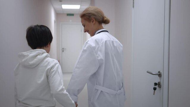 Positive Caucasian Pediatrician Talking To Patient Walking In Slow Motion To Office Along Hospital Corridor Closing Door. Cheerful Woman And Boy Holding Hands Strolling In Medical Clinic Indoors