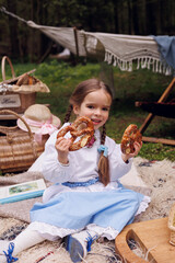 Little 5-eyar-old girl sitting on picnic in park and eating 
pretzel and buns. Little baby girl of 5-years-old in retro vintage dress having fun and smiling in forest on blanket