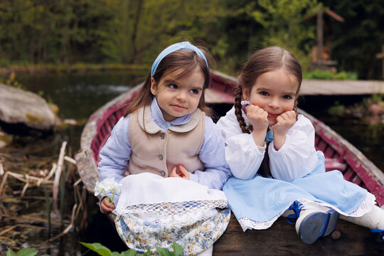 Two little girls of 3 and 5 sitting in old wooden pink boat in village. Little baby sisters in retro vintage dress smiling and thoughtful
