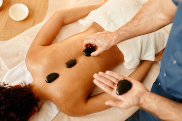 Top view closeup of male therapist laying stones on back of young woman in relaxing SPA session, copy space