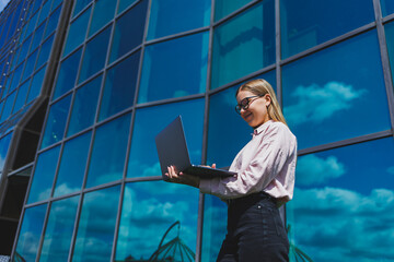 Concentrated cheerful woman in casual clothes and trendy glasses stands against the backdrop of a skyscraper with a laptop in her hands, smiles and surfs the internet on a laptop