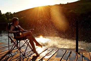 The girl is sitting on a chair by the lake. Children rest on the pier. Water splashes on sunset...