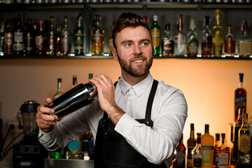 handsome barman in white shirt and black apron holds in hands steel shaker. Blurred background