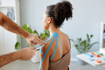 Close up rehabilitation therapist putting k tape on shoulder of young African American woman in physiotherapy clinic - obrazy, fototapety, plakaty