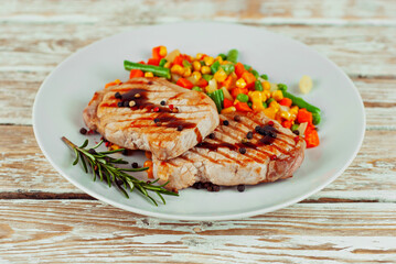 Pieces of meat grilled. Steak with rosemary and pepper in a white plate. Meat dish with red sauce on a white wooden table. Salad of corn, carrots and green peas near the steak.