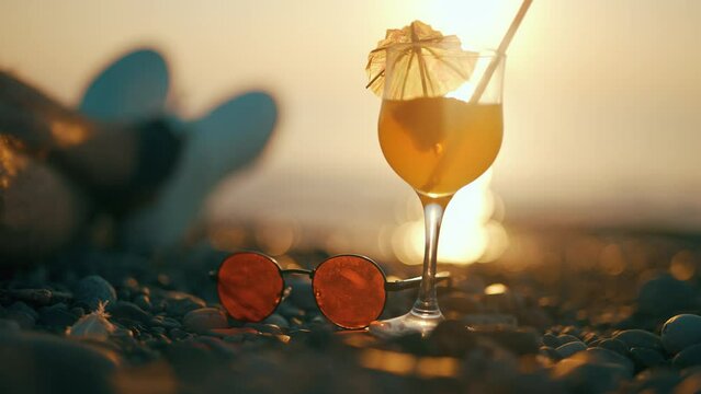 A man is enjoying summer sunset on background of a sea. Cool tropical drink and sunglasses with seashore. Waves are crashing on beach. Concept of travel vacation or relax and holidays. Close up.