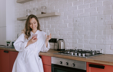 A young beautiful caucasian happy smiling blonde woman in a white robe with a glass of water in the morning checks messages in her mobile phone's mail on background of the kitchen