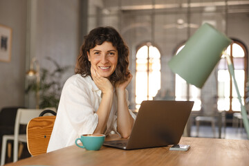 Beautiful young caucasian woman looks into camera sitting at table with laptop in cafe. Brunette...