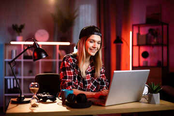 Focused caucasian woman working on modern laptop during evening time at home. Young lady hipster wearing checkered shirt and black cap. Concept of people, freelance and deadline.