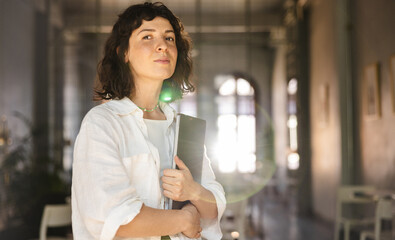 Young caucasian girl looks into camera holding laptop after online class in cafe. Brunette wearing white top, shirt. Concept of education