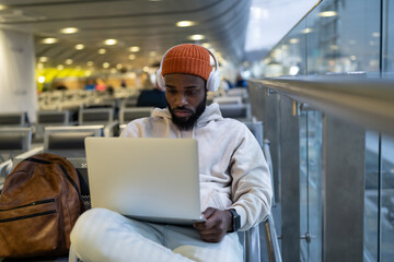 Young African American man sitting in hall of airport terminal using laptop, wear headphones and listening music. Passenger travelling with backpack, online working on computer and waiting landing. 