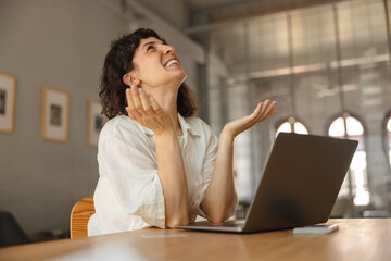 Pretty young european girl looks up waving her arms sitting at table in cafe. Brunette girl is happy with finished work on project. Concept of technology