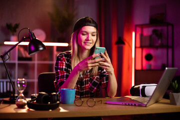 Naklejka na ściany i meble Pretty young woman in checkered shirt sitting at desk with modern smartphone in hands. Beautiful happy lady surfing internet on cell phone during evening time at home.