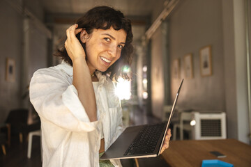 Pretty young caucasian woman looks into camera, tucking hair behind ear holding laptop. Brunette wears white shirt standing indoors. Concept of technological device.