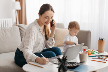 Happy business mother working at home while her little son playing next to her, lady talking on...