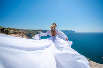 Blonde with long hair on a sunny seashore in a white flowing dress, rear view, silk fabric waving in the wind. Against the backdrop of the blue sky and mountains on the seashore.