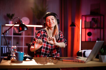 Portrait of caucasian woman wearing wireless headphones for listening music during evening time. Charming lady in cap smiling with closed eyes at home.