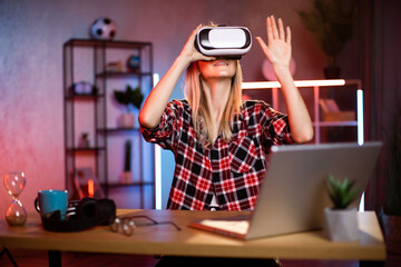 Young cheerful woman in VR goggles working in augmented reality while staying at home. Attractive female in red shirt sitting at desk with laptop and tapping on virtual screen.