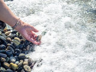 A hand with a bracelet made of stones holds a pebble near the water. Sea wave and female hand. Feel the wave.