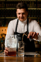 male bartender holds jigger and carefully pours drink into transparent glass with ice cubes.